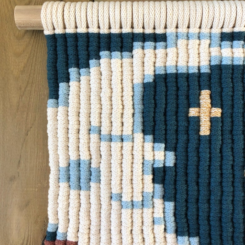 Pode incluir: Uma tape&ccedil;aria de parede feita de fio tecido em tons de creme, azul e azul-petr&oacute;leo. O fio &eacute; tecido em um padr&atilde;o geom&eacute;trico com um sinal de mais dourado no centro.