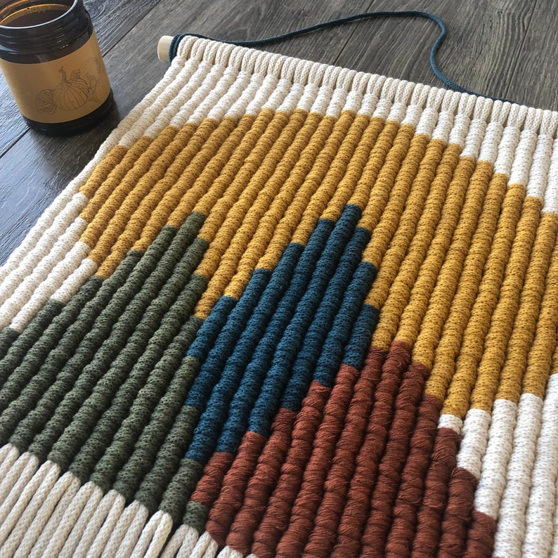 May include: A wall hanging made of woven yarn in a geometric pattern. The colors are white, mustard yellow, dark green, navy blue, and brown.