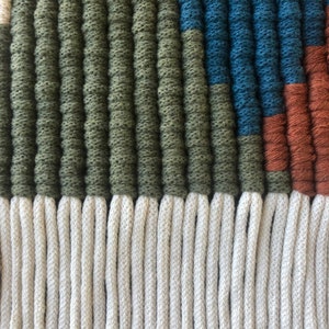 May include: Close-up of a woven wall hanging with a geometric pattern in shades of green, blue, and brown. The bottom of the hanging features a fringe of white yarn.