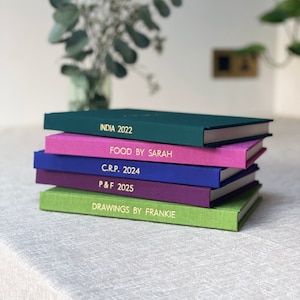 Puede incluir: Una pila de diarios de tapa dura de colores, con letras doradas. Los diarios son de color verde, rosa, azul, morado y verde claro. Los títulos incluyen "INDIA 2022", "FOOD BY SARAH", "C.R.P. 2024", "P&F 2025" y "DRAWINGS BY FRANKIE".