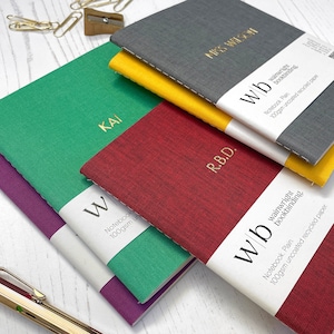 May include: Five notebooks with personalized names and the text "Wainwright Bookbinding" on the covers. The notebooks are in various colors, including red, green, purple, gray, and yellow. The notebooks are made of 100gsm uncoated recycled paper.