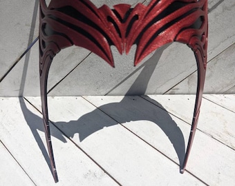 May include: A metallic red headpiece featuring a pointed, symmetrical design with intricate cutouts and raised ridges. The accessory has a wide, curved front section that tapers into two thin, vertical side bands, creating a distinct crown-like shape.