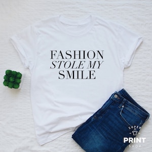 May include: White t-shirt with black text that reads "FASHION STOLE MY SMILE". The shirt is laid flat on a white surface with a pair of blue jeans and a small potted plant.