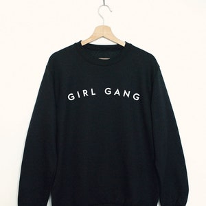 Può includere: Felpa nera con la scritta "GIRL GANG" stampata in bianco sul davanti.