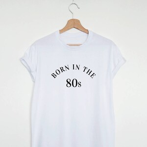 May include: A white t-shirt with the text "BORN IN THE 80s" in black, displayed on a wooden hanger. The shirt has a classic crew neck and rolled-up sleeves, suggesting a casual style.