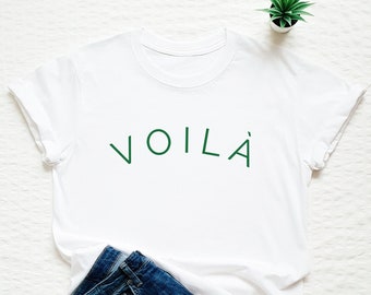 May include: White t-shirt with green text that says "VOILA" on it. The shirt is paired with blue jeans and a small potted plant.