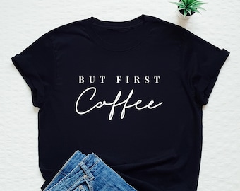 May include: A black t-shirt with the white text "BUT FIRST Coffee" in a mix of serif and script fonts. The shirt is paired with blue denim jeans. A small potted plant is visible in the background.
