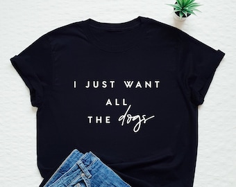 May include: A black t-shirt with white text that reads "I just want all the dogs". The shirt is laying on a white surface with a pair of blue jeans.