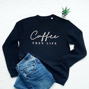 May include: A black sweatshirt with the text "Coffee then life" in white cursive lettering. The sweatshirt is folded and laid on a white surface with a pair of blue jeans.