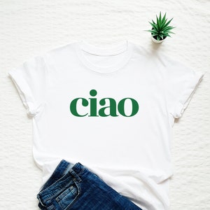 Pode incluir: Camiseta branca com uma estampa gr&aacute;fica verde de "ciao". A camiseta est&aacute; dobrada e colocada sobre uma superf&iacute;cie branca com um par de cal&ccedil;as de ganga azuis e uma pequena planta em vaso.