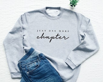 May include: A grey sweatshirt with the text "JUST ONE MORE chapter" in black lettering. The sweatshirt is laid flat on a white surface with a pair of blue jeans.