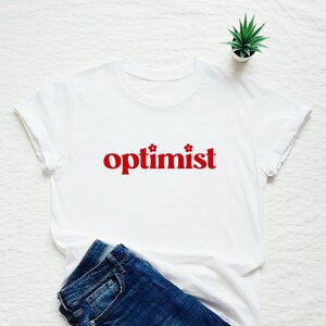 Op de afbeelding: Wit T-shirt met rode tekst "optimist" met twee kleine rode bloemen aan weerszijden van het woord.