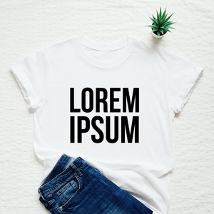 Peut inclure: T-shirt blanc avec un texte noir qui dit "LOREM IPSUM". Le t-shirt est plié et posé sur une surface blanche avec un jean bleu et une petite plante en pot.
