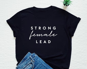 May include: Black t-shirt with white text that reads "STRONG female LEAD". The shirt is laid flat on a white surface with a pair of blue jeans folded next to it.