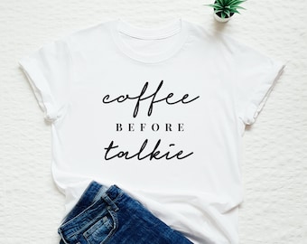 May include: White t-shirt with black text that reads "coffee before talkie". The shirt is lying on a white surface with a pair of blue jeans and a small potted plant.