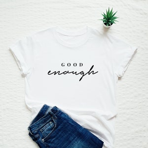 May include: White t-shirt with the text "GOOD enough" printed in black on the front. The shirt is laid flat on a white surface with a pair of blue jeans and a small potted plant.