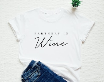 May include: White t-shirt with the text "Partners in Wine" in black script font. The shirt is laid flat on a white surface with a pair of blue jeans folded next to it.