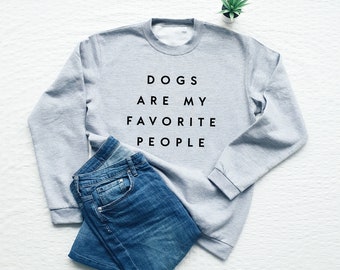 May include: A gray sweatshirt with the text "Dogs are my favorite people" printed on the front. The sweatshirt is laid flat on a white surface with a pair of blue jeans folded next to it.