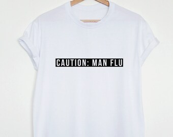 May include: White t-shirt hanging on a wooden hanger. The shirt has the text "CAUTION: MAN FLU" printed in black, centered horizontally. The shirt's sleeves are rolled up.
