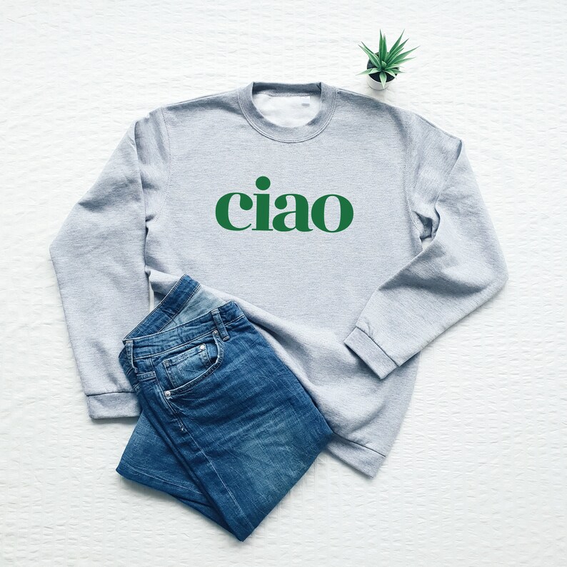 May include: A gray sweatshirt with the word "ciao" printed in green on the front. The sweatshirt is laid out on a white surface with a pair of blue jeans and a small potted plant.