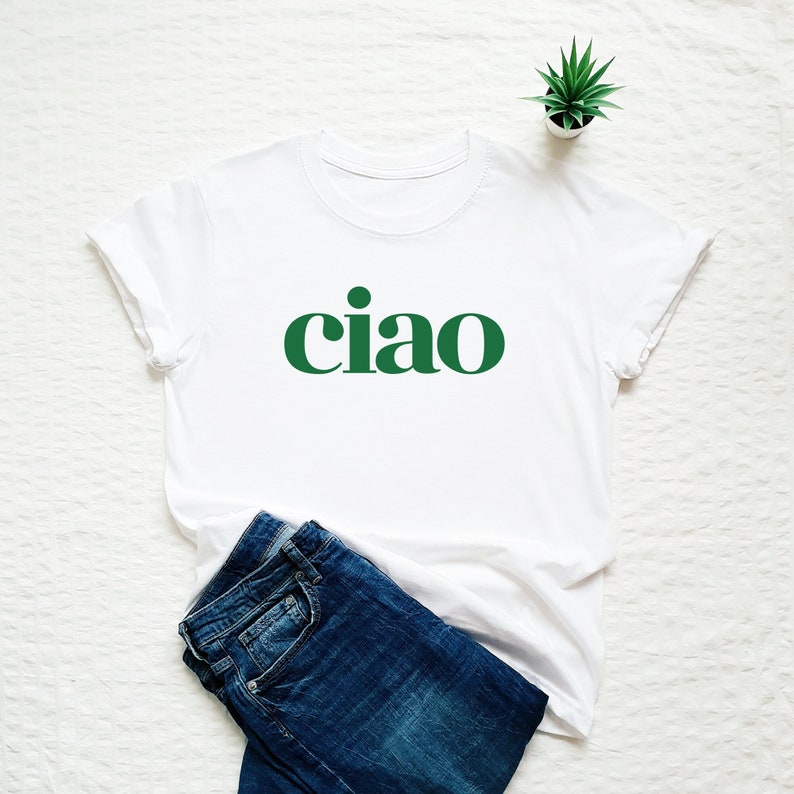 Pode incluir: Camiseta branca com uma estampa gr&aacute;fica verde de "ciao". A camiseta est&aacute; dobrada e colocada sobre uma superf&iacute;cie branca com um par de cal&ccedil;as de ganga azuis e uma pequena planta em vaso.