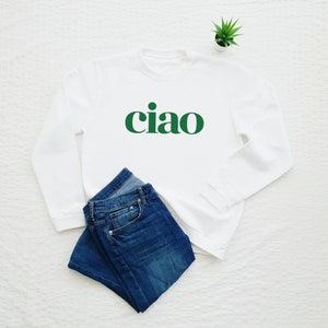 May include: White sweatshirt with long sleeves and a green "ciao" graphic print. The sweatshirt is laid flat on a white surface with a pair of blue jeans folded next to it.