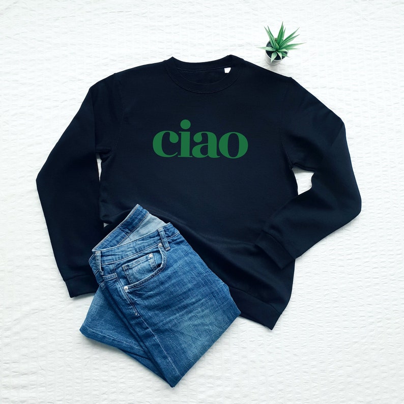 May include: A dark blue sweatshirt with a green "ciao" graphic print. The sweatshirt is folded and laid on a white surface with a pair of blue jeans.