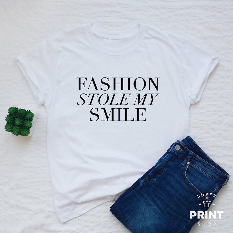 May include: White t-shirt with black text that reads "FASHION STOLE MY SMILE". The shirt is laid flat on a white surface with a pair of blue jeans and a small potted plant.
