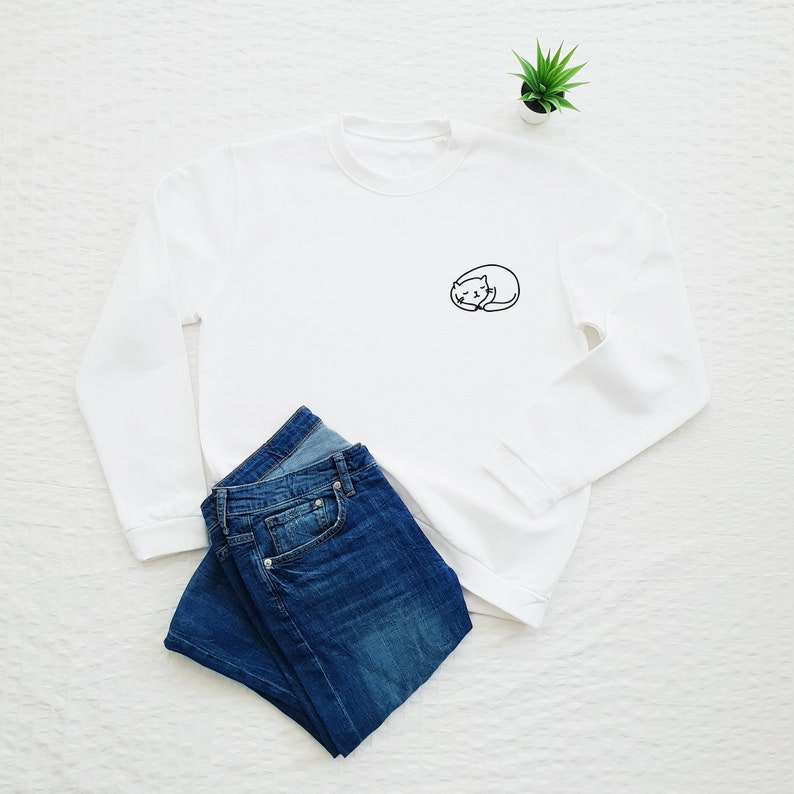 Puede incluir: Sudadera blanca de manga larga con una peque&ntilde;a imagen bordada de un gato durmiendo en el pecho izquierdo. La sudadera est&aacute; colocada sobre una superficie blanca con un par de pantalones vaqueros azules doblados encima.