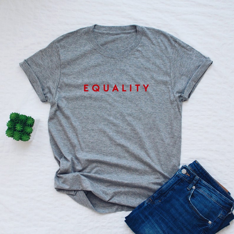 May include: A gray t-shirt with the word "EQUALITY" printed in red on the front. The shirt is folded and laid on a white surface with a pair of blue jeans.