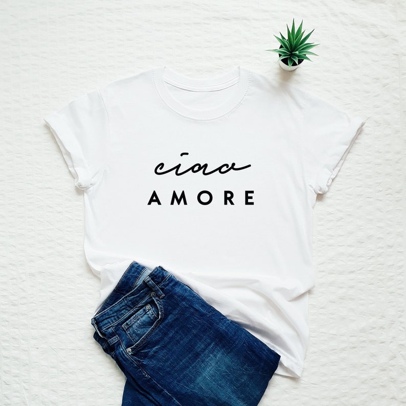Op de afbeelding: Wit T-shirt met zwarte tekst die "ciao amore" luidt op een witte achtergrond. Het shirt is gevouwen en ligt op een wit oppervlak met een blauwe spijkerbroek en een kleine plant in een pot.