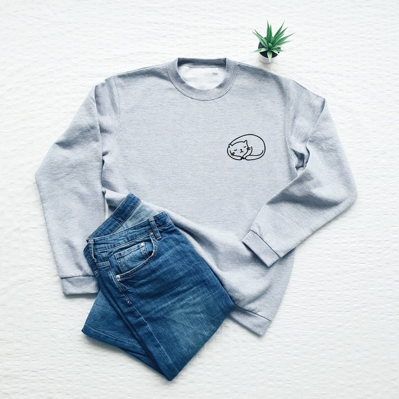 Puede incluir: Una sudadera gris con un peque&ntilde;o dise&ntilde;o de gato bordado en el pecho izquierdo. La sudadera se combina con unos vaqueros azules.