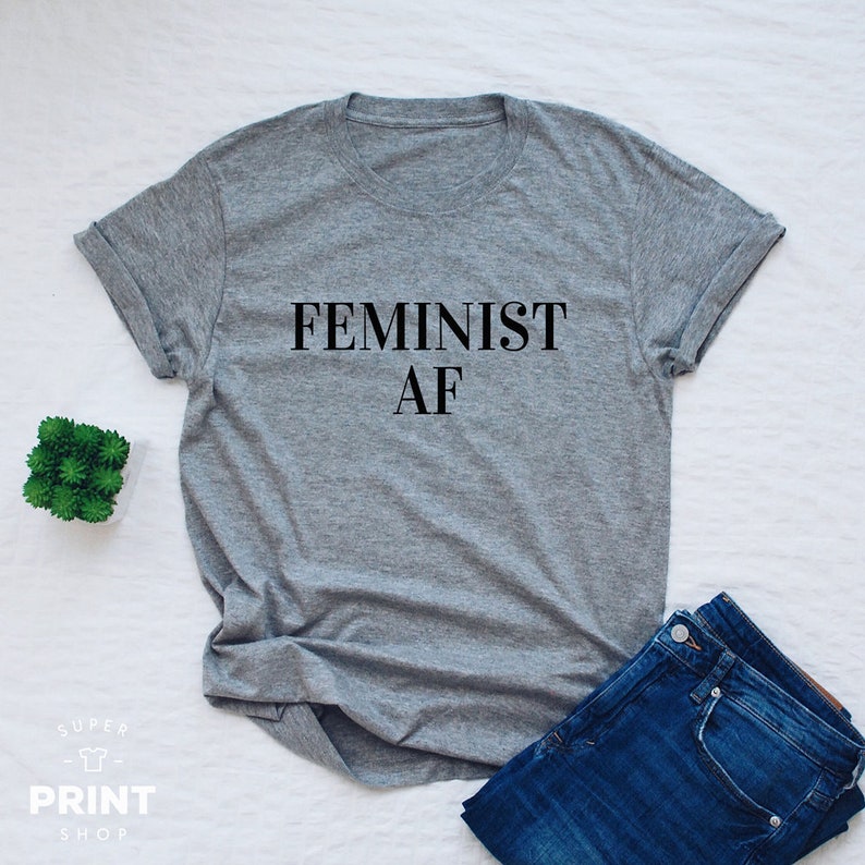 Puede incluir: Una camiseta gris con el texto "FEMINIST AF" impreso en negro.