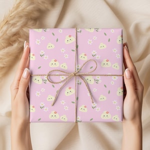 May include: A square gift box wrapped in light pink paper featuring a repeating pattern of small yellow cartoon dumplings with smiling faces, white daisy flowers, and green leaves, tied with a thin brown twine string in a center bow.
