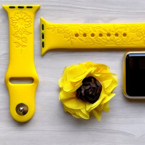 May include: A yellow silicone Apple Watch band with a sunflower design. The band is shown with a matching yellow watch face and a yellow fabric sunflower.
