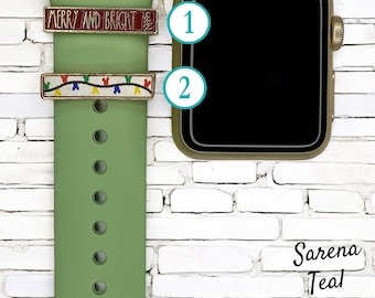 Puede incluir: Dos dijes decorativos para correas de reloj. Un dije tiene las palabras "MERRY AND BRIGHT" sobre un fondo rojo. El otro dije tiene una cadena de luces de colores con cabezas de Mickey Mouse. Ambos dijes son dorados y están sujetos a una correa de reloj verde.