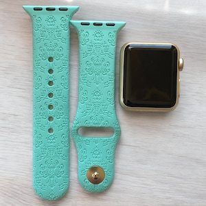 May include: Two teal blue Apple Watch bands with a Haunted Mansion pattern. The bands are made of silicone and have a gold buckle. The bands are shown next to a gold Apple Watch.