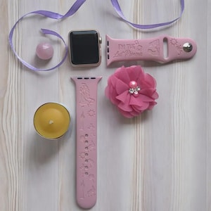 May include: A light pink silicone watch band featuring engraved icons and the text I'M THE LOST PRINCESS, paired with a matching watch face, a pink fabric flower accessory with central beads, a small round pink bead, and a yellow tea light candle.