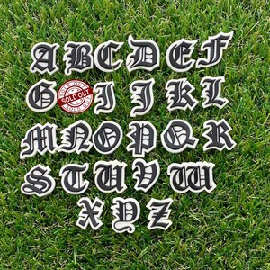 以下が含まれることがあります： ゴシック体で書かれた、黒と白の26個の大文字のセット。アルファベット順に並べられており、緑の芝生の背景に置かれています。Gの文字には赤い「完売」のスタンプが押されています。