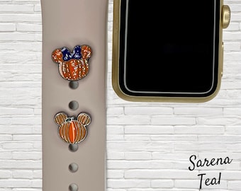 Könnte beinhalten: Ein rosafarbenes Apple Watch-Armband mit zwei Kürbis-Mickey-Mouse-Anhängern. Die Anhänger sind orange und schwarz mit Glitzerakzenten. Das Armband ist auf einem weißen Ziegelhintergrund. Der Text "Sarena Teal Designs" ist in der unteren rechten Ecke des Bildes sichtbar.
