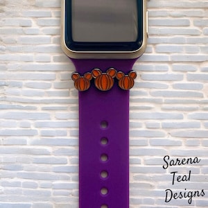 May include: A purple watch band with three orange pumpkin charms attached. The watch band is connected to a gold watch. The background is a white brick wall. The text "Sarena Teal Designs" is in the bottom right corner.