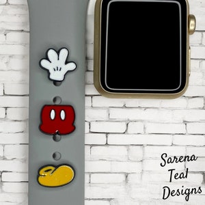 May include: A gray Apple Watch band with three Mickey Mouse-themed charms: a white glove, red shorts, and a yellow shoe. The charms are attached to the band with small holes. The band is on a white brick wall. The text "Sarena Teal Designs" is visible in the lower right corner of the image.