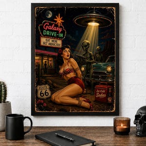 May include: A framed wall art print featuring a retro sci-fi scene with a person in a red polka dot outfit kneeling before an alien holding a ray gun. A UFO hovers above a vintage car near a Galaxy Drive-In sign that reads Eat Here Get Abducted.