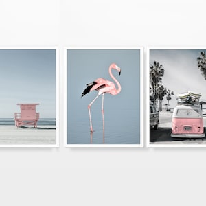 May include: Set of three framed prints featuring beach scenes. One shows a pink lifeguard stand on a sandy beach, another a pink flamingo in water, and the third a pink van with surfboards, palm trees in the background.