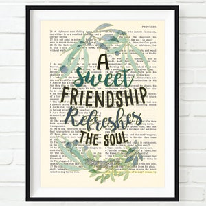 May include: A framed print featuring the words "A sweet friendship refreshes the soul" in a decorative font, surrounded by a wreath of olive branches. The background is a vintage book page.