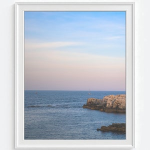 Pu&ograve; includere: Una fotografia incorniciata di un paesaggio marino con un cielo blu e una costa rocciosa. L'acqua &egrave; di un blu profondo e il cielo &egrave; di un azzurro chiaro con un tocco di rosa. Ci sono due piccole barche in lontananza.