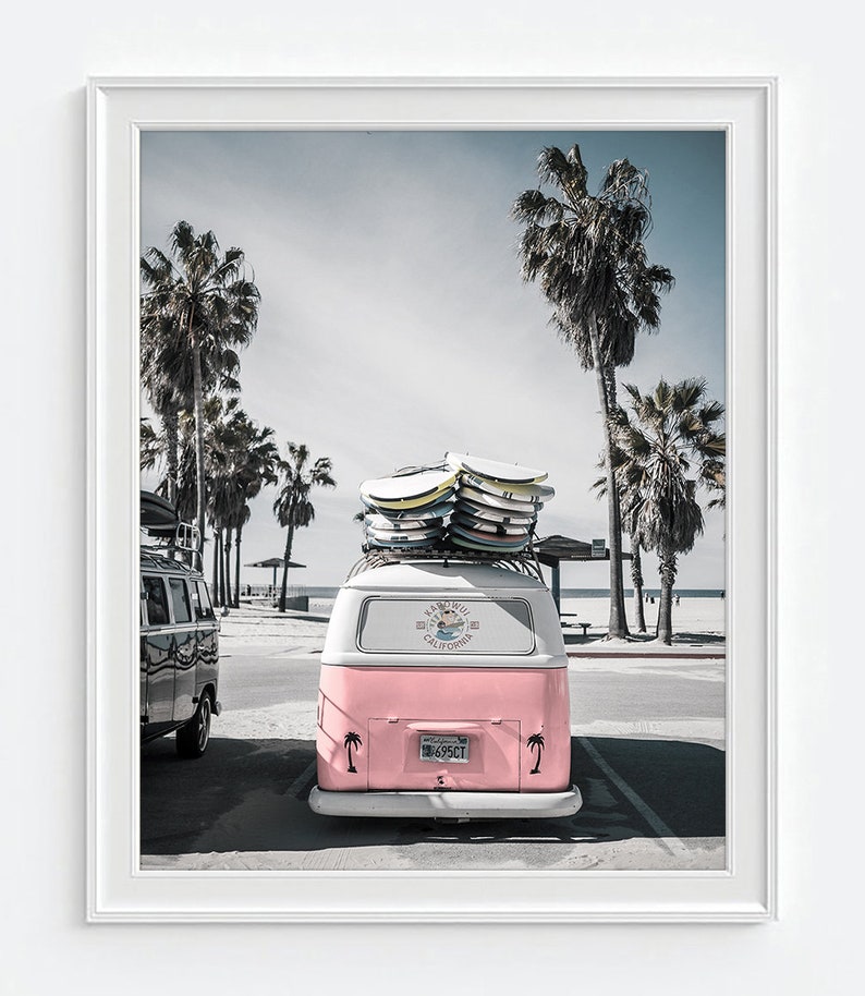 May include: A framed print featuring a pink and white vintage van parked on a beach, topped with surfboards. Palm trees line the beach and the van has a California license plate. The sky is a pale blue.