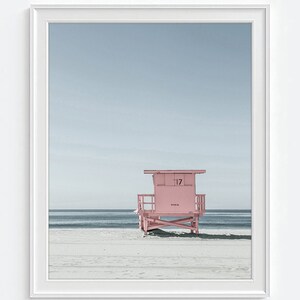 May include: A framed print featuring a pink lifeguard stand on a sandy beach under a pale blue sky. The number 17 is visible on the stand. The ocean is in the background.
