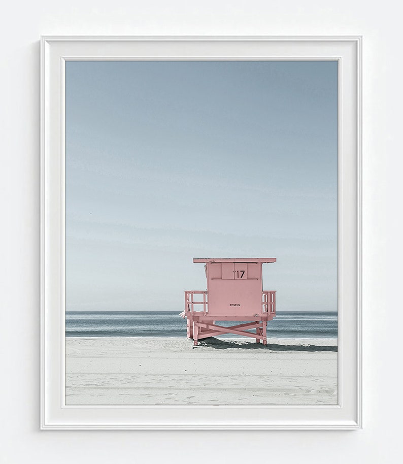 May include: A framed print featuring a pink lifeguard stand on a sandy beach under a pale blue sky. The number 17 is visible on the stand. The ocean is in the background.