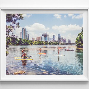 K&ouml;nnte beinhalten: Ein Panoramablick auf eine Skyline mit mehreren Geb&auml;uden, darunter ein hoher Wolkenkratzer, die sich im Wasser spiegeln. Menschen paddeln auf dem Wasser vor der Stadt.
